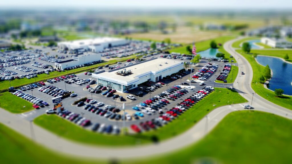 Aerial high-angle view of a bustling car dealership surrounded by parked cars in a green landscape.
