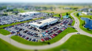 Aerial high-angle view of a bustling car dealership surrounded by parked cars in a green landscape.